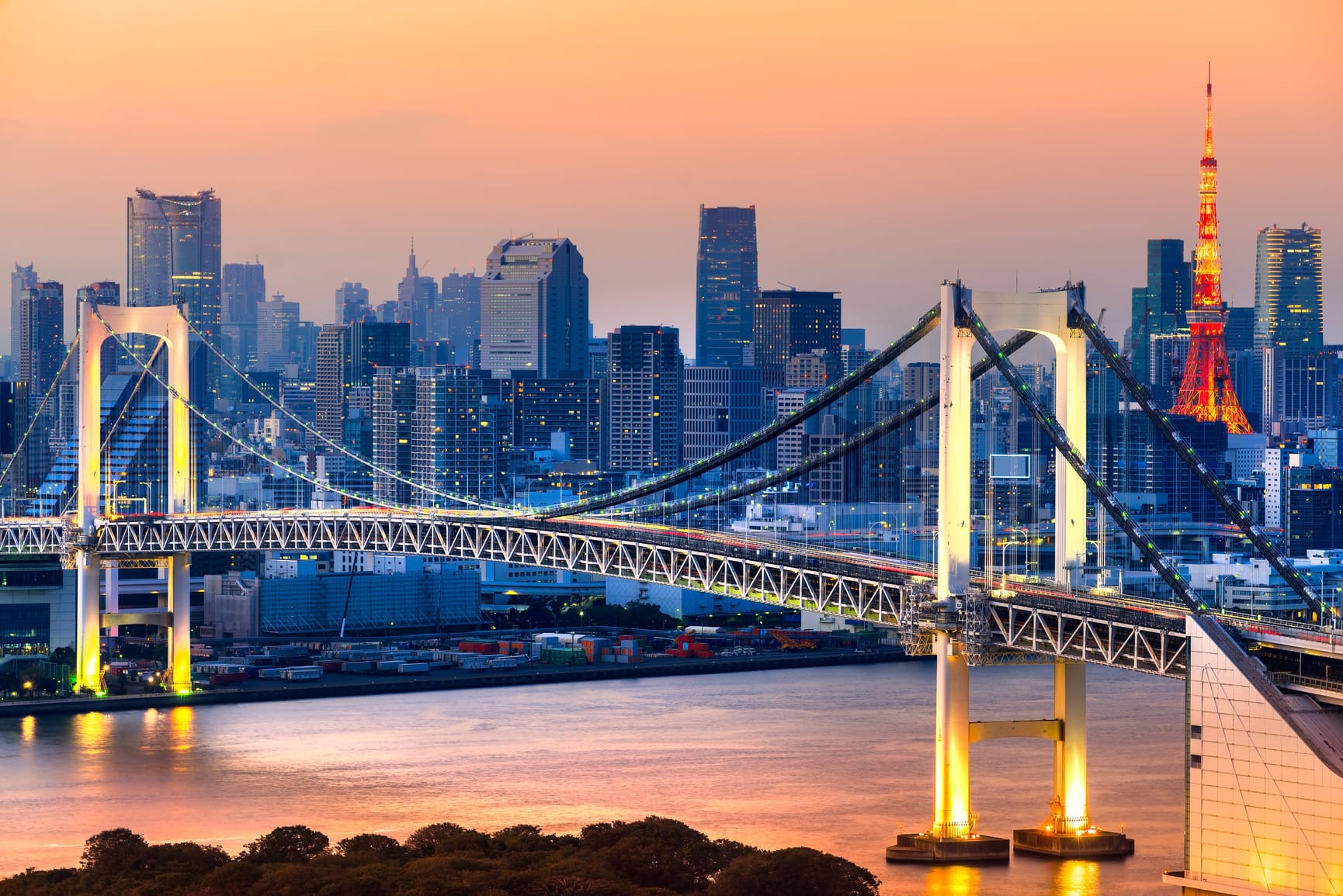 Sunset image of rainbow bridge in Tokyo, with skyline in background and river below.