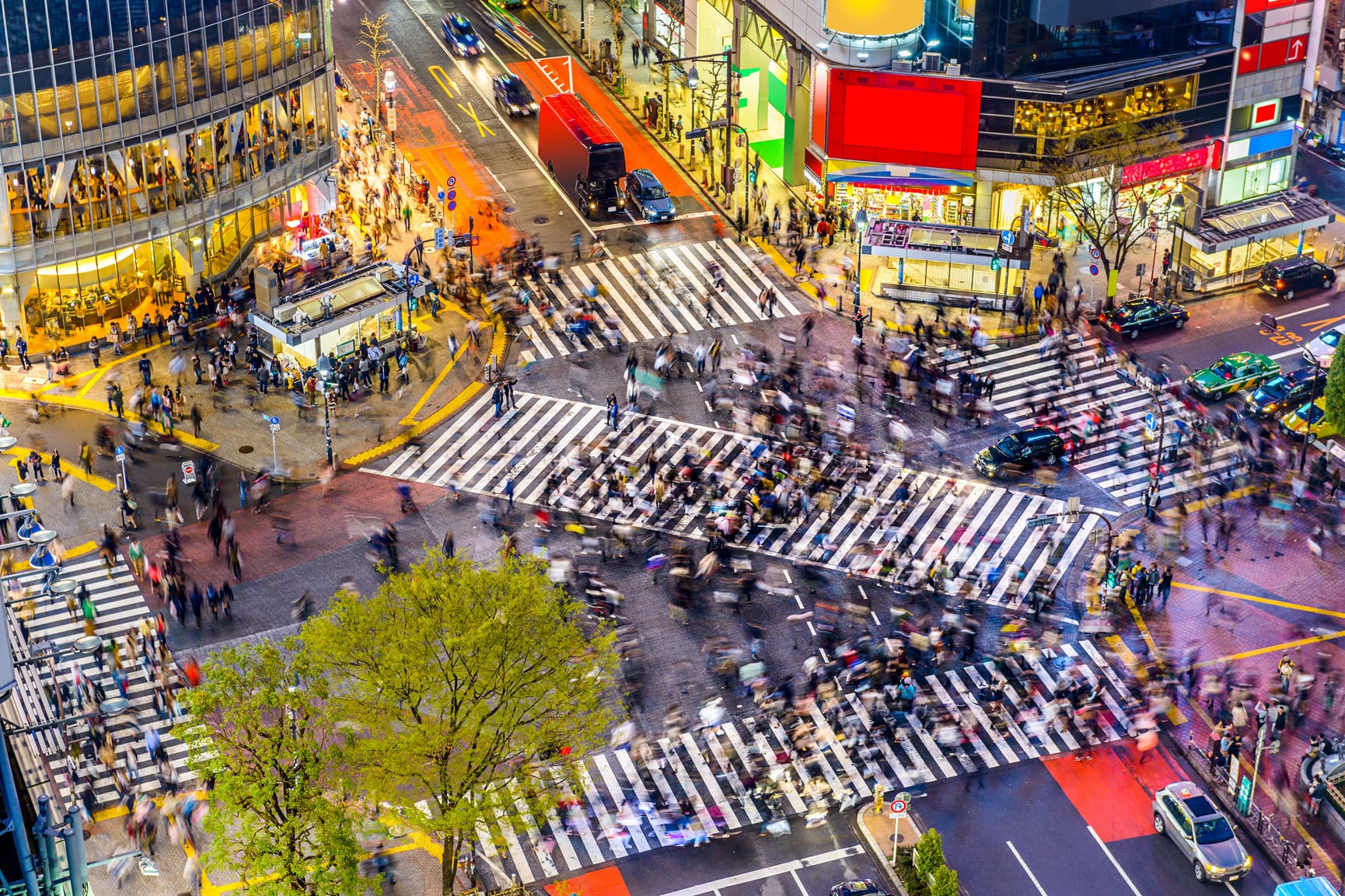 Overhead shot of intersection of Shibuya with buildings lit up and motion blur of people crossing intersection.